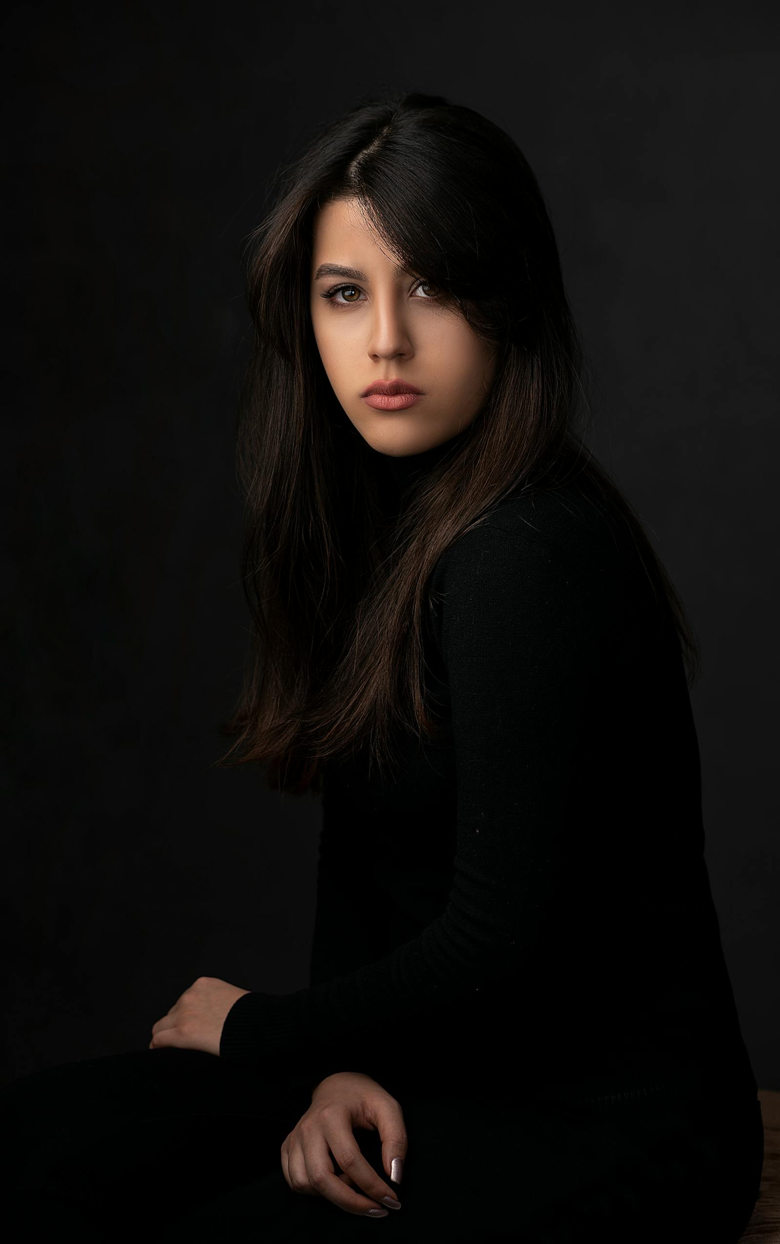 A studio portrait lit in warm tungsten — a young woman in profile against a deep charcoal seamless, soft fall-off on her shoulder, signature Mira Beauty lighting.